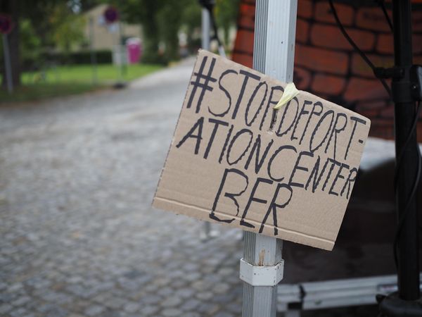 An einem Pavillon hängt ein Pappschild mit der Aufschrift "#StopdeportationairportcenterBER"