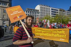 Im Vordergrund steht ein Mann der ein Oranges Schild mit der Aufschrift "Solidarisch warm statt sozial kalt" hält, im Hintergrund ist eine Kundgebung zu sehen und ein ebenfalls oranges Transparent mit dem Slogan "Energiewirtschaft vergesellschaften!"