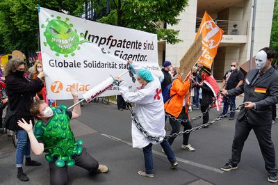 Eine Ärztin wird von einem Menschen im Anzug mit Deutschland-Fahne und Biontech-Logo an Ketten gehalten, aber schafft es dennoch gerade so einen Menschen im Coronakostüm mit einer Impfspritze zu stechen und zu Fall zu bringen