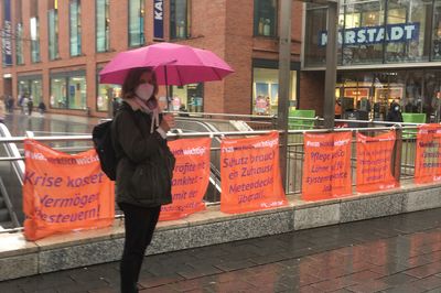 Am U-Bahneingang sind mehrere Transparente für Gesundheit und Solidarität in der Corona-Zeit angebracht, davor steht eine Aktivistin mit Regenschirm