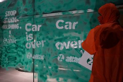 Auf einem  grün angesprühten Schaufenster steht weiß abgesprüht "Car is over – attac", davor steht ein Mensch im orangen Overall mit einer Sprühschablone in der Hand 