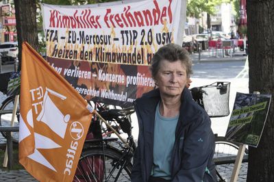 Eine Frau mit einer orangen Attac-Fahne bei der ein Mensch die Buchstaben "CETA" in eine Mülltonne schmeißt steht vor einem Transparent mit der Aufschrift "Klimakiller Freihandel"