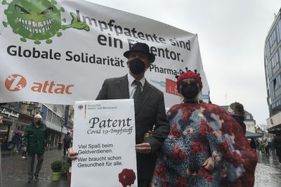 Eine Person im Coronakostüm und eine Person im Anzug, der ein Schild mit einem Impfpatent in der Hand hält stehen vor einem Banner mit dem Slogan "Impfpatente sind ein Eigentor."