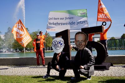 Wissing und die Autoindustrie sitzen im Gras für den brennenden Resten der 1,5°, im Hintergrund halten zwei Aktivist*innen ein Banner mit dem Slogan "Don't burn our future: Verkehrswende jetzt!"