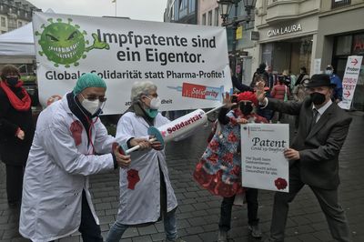 Zwei als Arzt:innen verkleidete Menschen halten eine Spritze mit der Aufschrift "Solidarität" in der Hand und richten sie gegen eine Person im Coronakostüm und eine Person im Anzug, der ein Schild mit einem Impfpatent in der Hand hält. Im Hintergrund ist ein Banner mit dem Slogan "Impfpatente sind ein Eigentor."