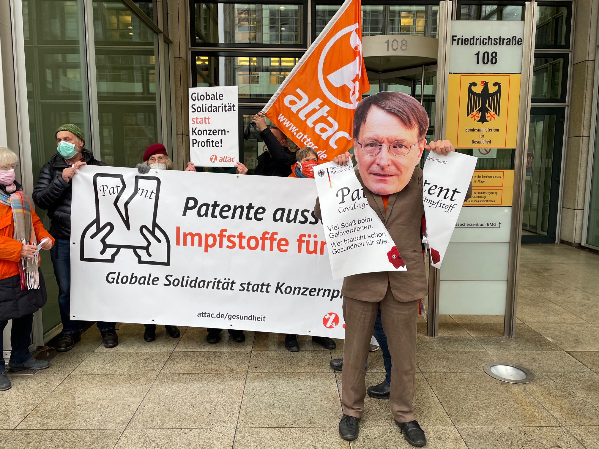 Ein als Karl Lauterbach verkleideter Aktivist zerreißt vor dem Bundesgesundheitsministerium ein übergroßes Patent. Im Hintergrund ist ein Transparent mit dem Slogan "Patente aussetzen – Impfstoffe für alle!"