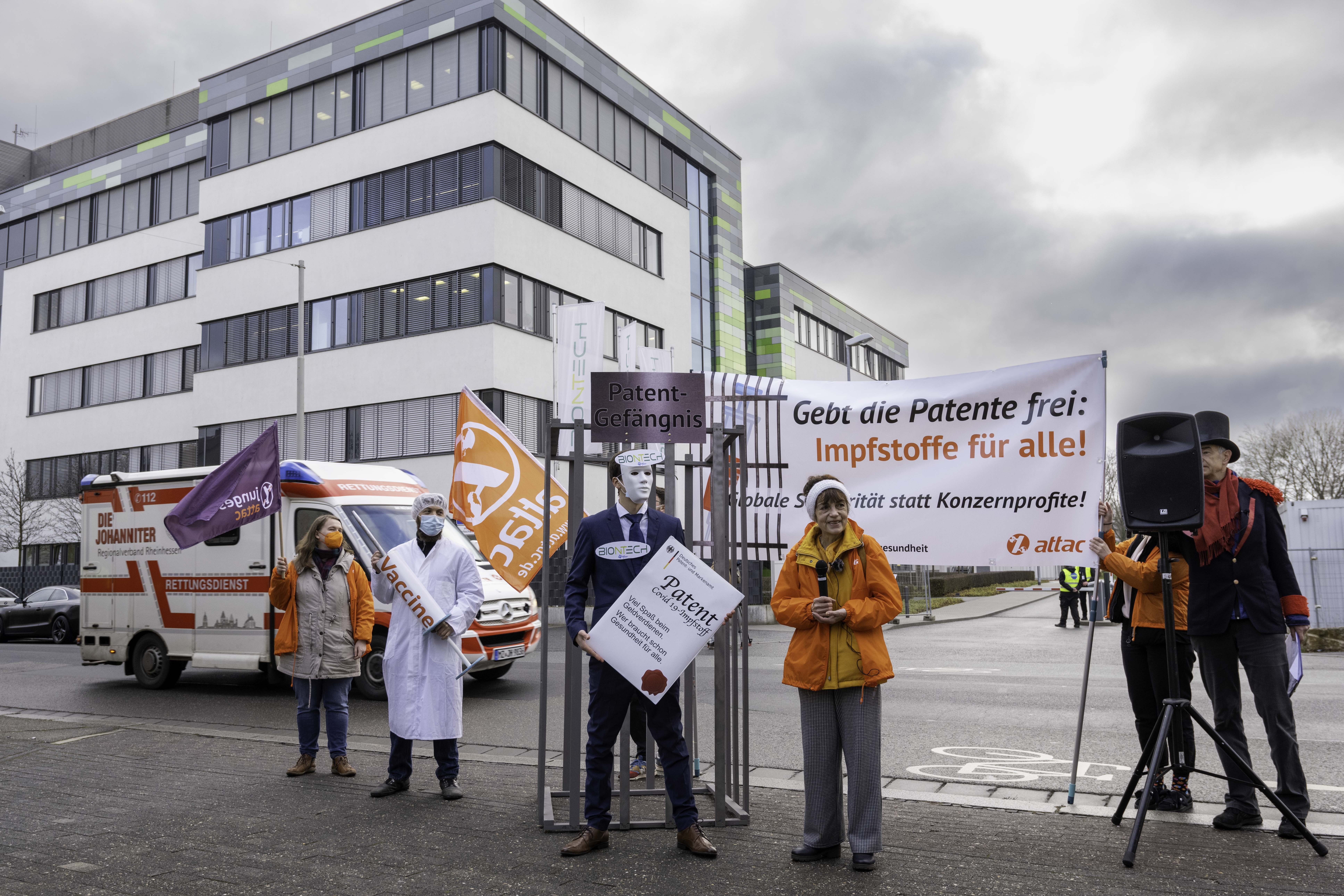 Ein Panoramabild er Protestierenden, links eine Aktivistin mit junges Attac-Flagge, dann ein Mensch der als Arzt verkleidet ist und eine übergroße Impfspritze hält, ein Patente-Käfig und ein Mann im Anzug der ein übergroßes Impfpatent hält.