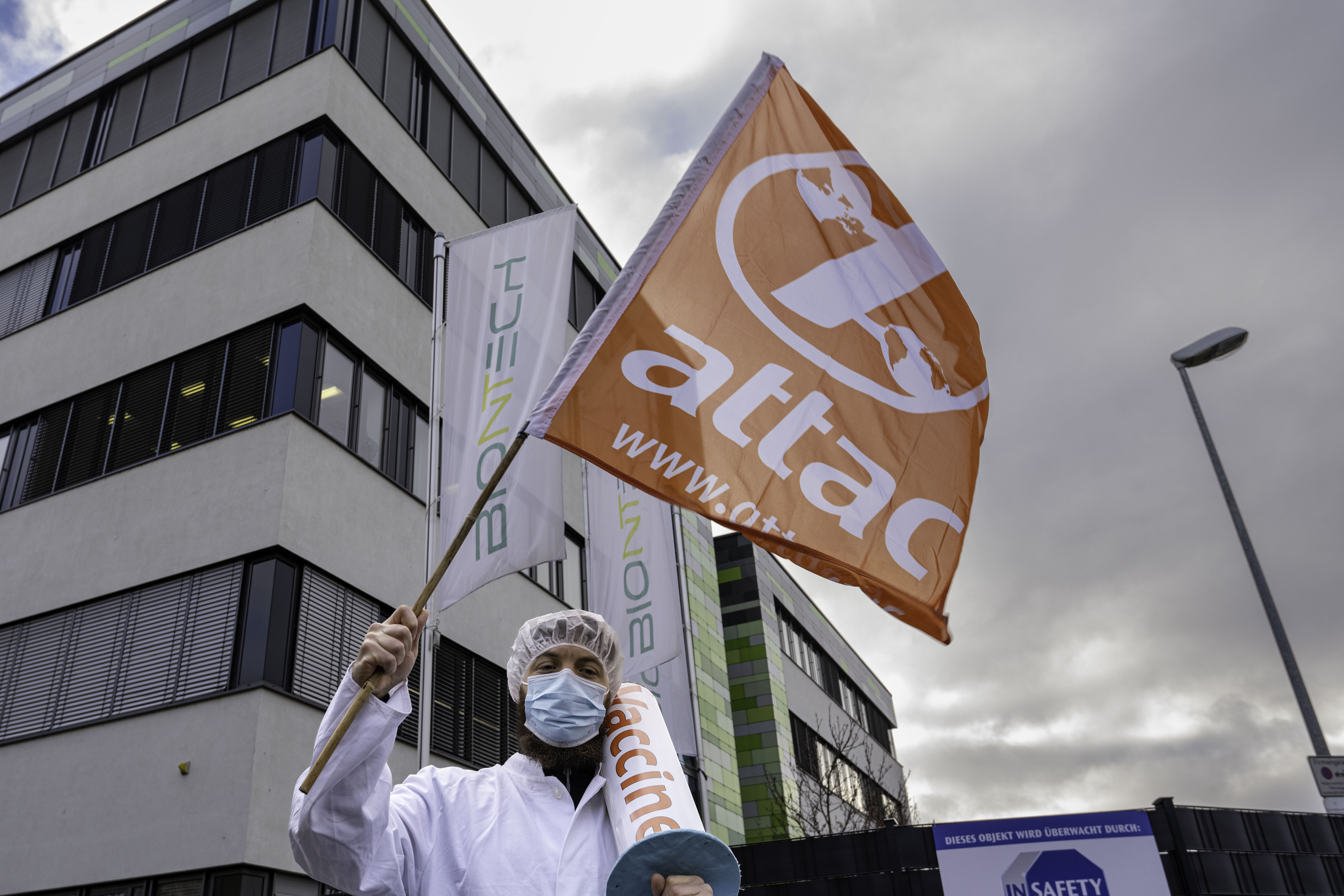 Der Impfarzt steht mit einer Attac-Fahne in der Hand triumphierend vor dem Biontech-Hauptgebäude