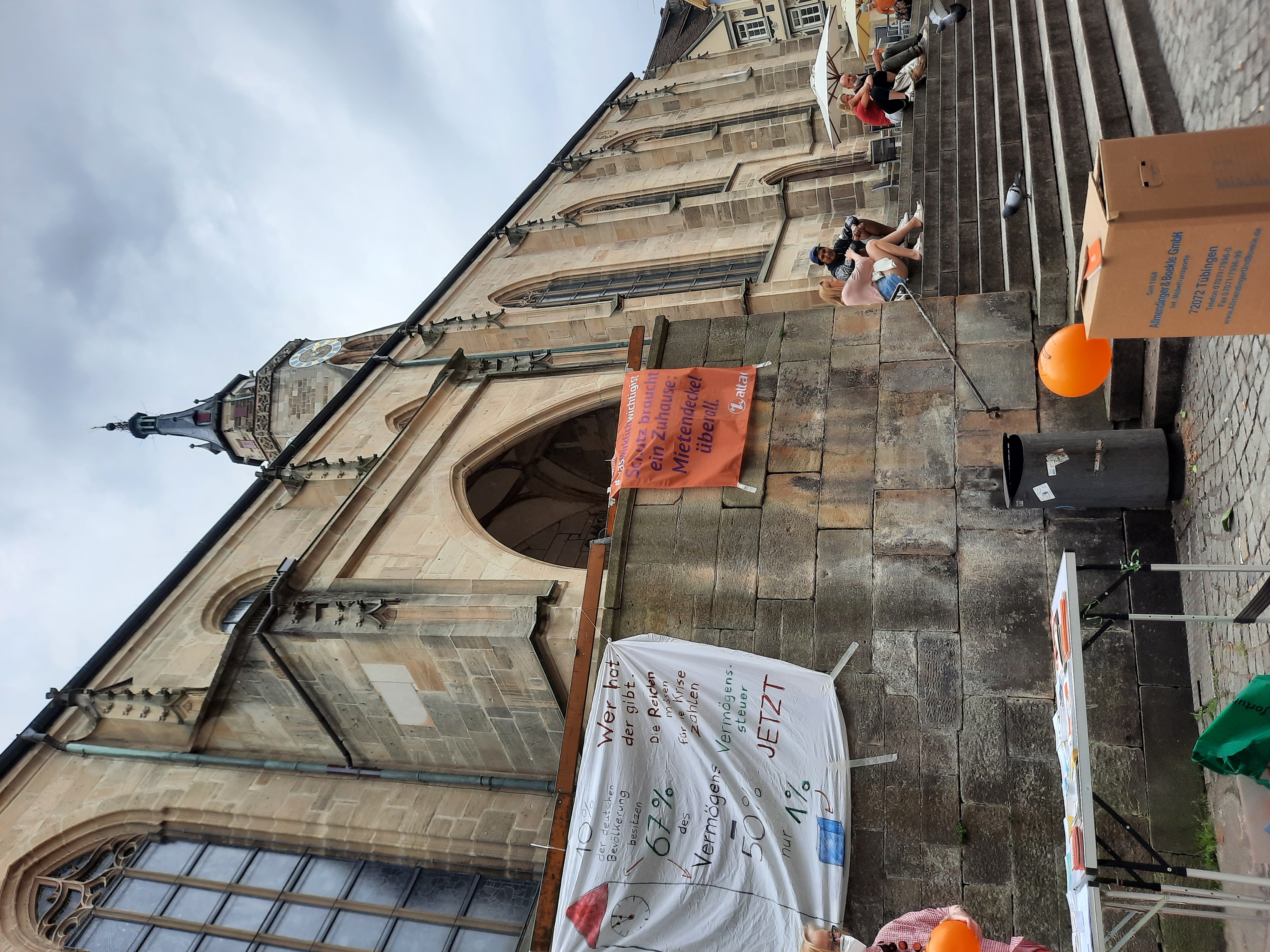 Im Bild ist ein Infostand von Attac in Tübingen, im Vordergrund ist ein Umzugskarton, der für das Vermögen der ärmeren Bevölkerungshälfte steht und im Hintergrund der Kirchturm der Marktkirche, die 67m Hoch ist und damit das Vermögen der reichsten 10% als Vergleich darstellt.
