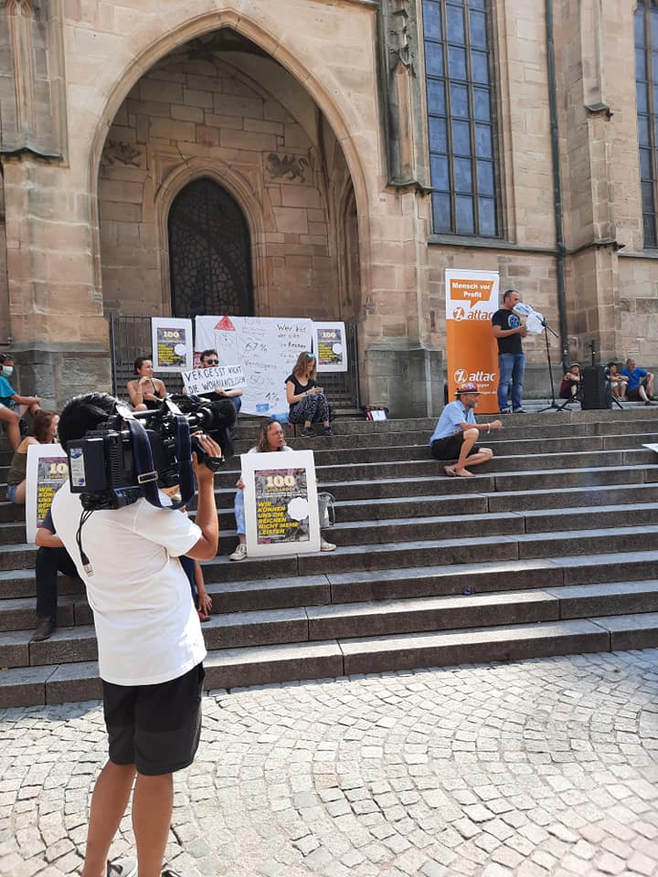 Auf den Stufen vor einer Kirche sitzen Menschen mit Transparenten und Plakaten von Wer hat der gibt, im Vordergrund ist ein Kameramann zu sehen