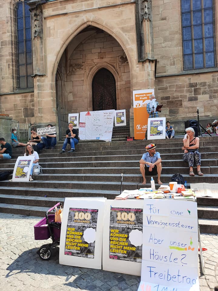Auf der Treppe vor einer Kirche sitzen Menschen für eine Kundgebung, im Vordergrund ist ein Infostand mit einem Plakat auf dem "Für eine Vermögenssteuer! Aber was ist mit unserem Häusle? Freibetrag 1 Mio. Euro" steht