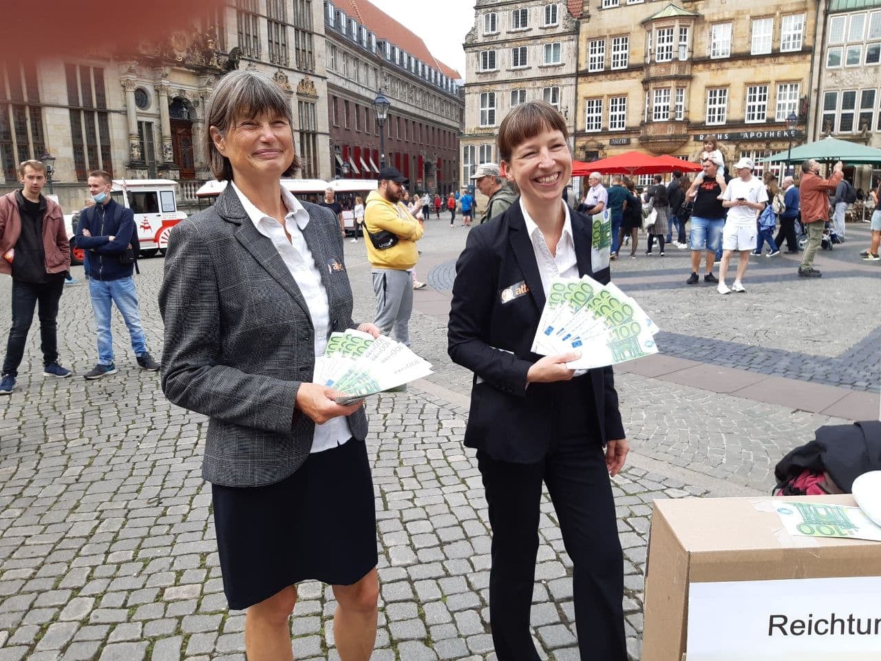 Zwei Frauen im Buiseneskostüm stehen auf dem Bremer Marktplatz und halten Flyer, die wie 100€ Scheine aussehen gefächert in ihrer Hand. 