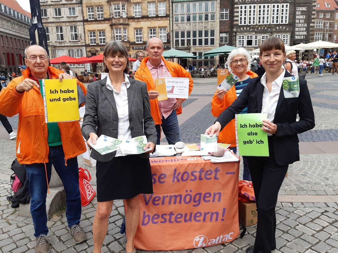 Bild von einem Infostand von Attac auf dem Marktplatz in Bremen, zu sehen sind Aktivist*innen in orangen Jacken und davor zwei Frauen im Buiseneskostüm mit 100€ Scheinen, dazu ein kleines Plakat auf dem "Ich habe, also gebe ich" steht.