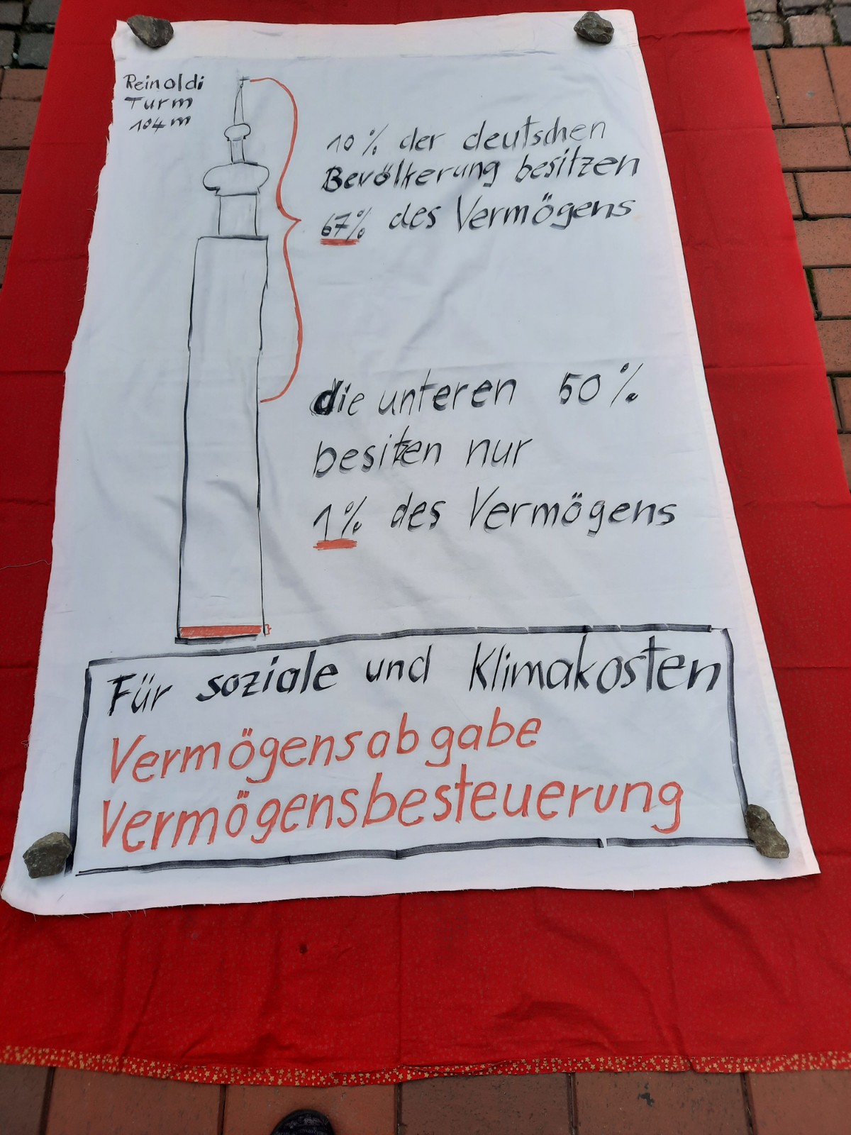 ein Plakat, das die Vermögensverteilung in Deutschland mit der Höher der dahinter liegenden Reinholdikirche Vergleicht und erklärt, dass die reichsten 10 Prozent 67 Prozent des Gesamtvermögens besitzen, während die ärmere Hälfte gerade ein Prozent besitzt.