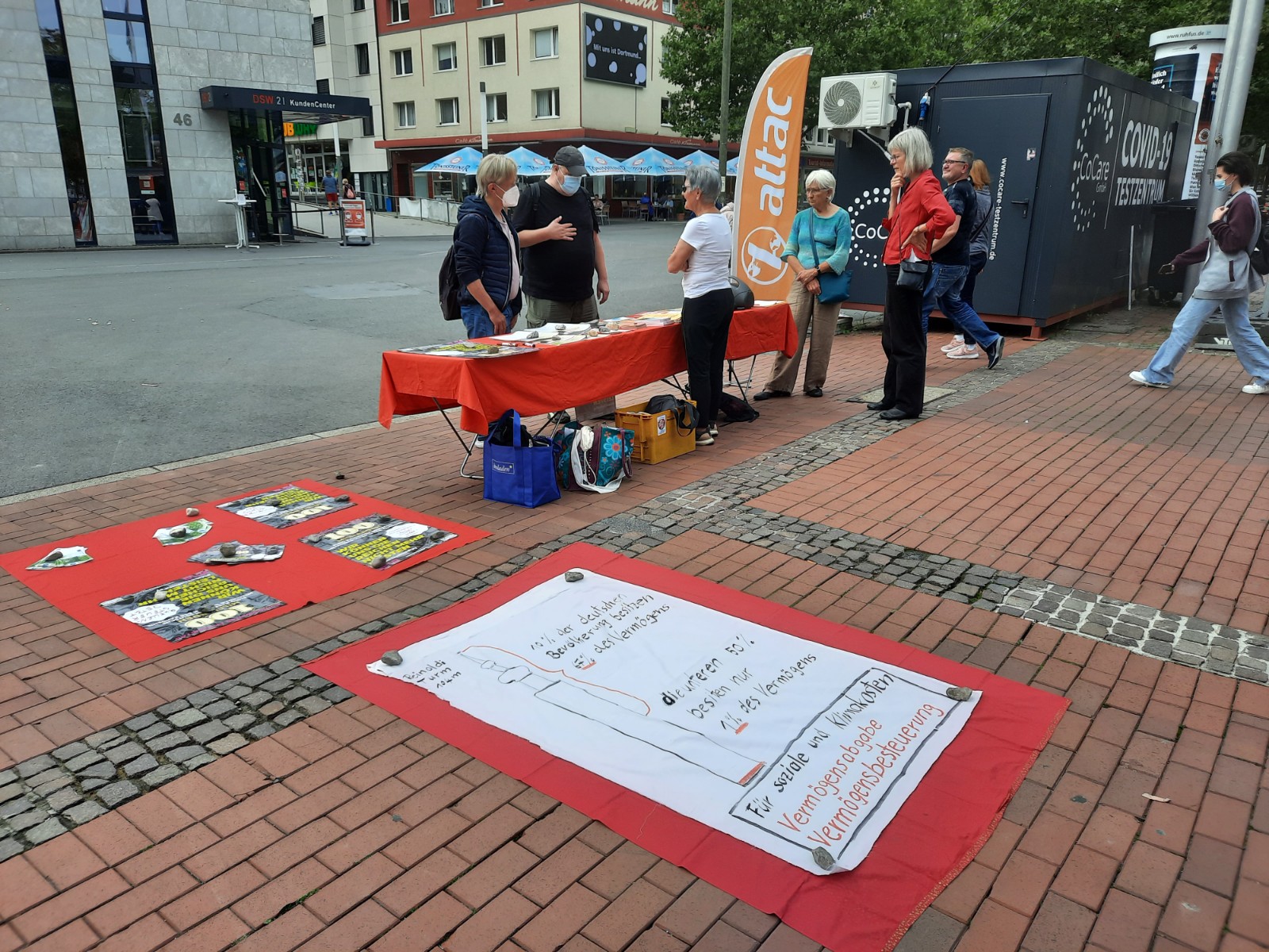 Im Bild ist ein Infostand mit Attacies, im Vordergrund ist ein Plakat, das die Vermögensverteilung in Deutschland mit der Höher der dahinter liegenden Reinholdikirche Vergleicht und erklärt, dass die reichsten 10 Prozent 67 Prozent des Gesamtvermögens besitzen, während die ärmere Hälfte gerade ein Prozent besitzt.