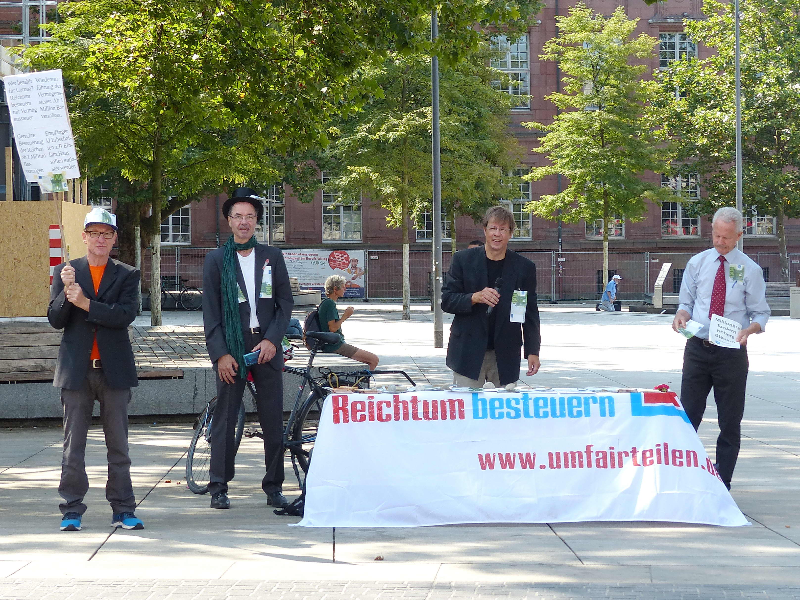 Im Bild ist ein Infostand mit Attacies, im Vordergrund ist ein Plakat, das die Vermögensverteilung in Deutschland mit der Höher der dahinter liegenden Reinholdikirche Vergleicht und erklärt, dass die reichsten 10 Prozent 67 Prozent des Gesamtvermögens besitzen, während die ärmere Hälfte gerade ein Prozent besitzt.