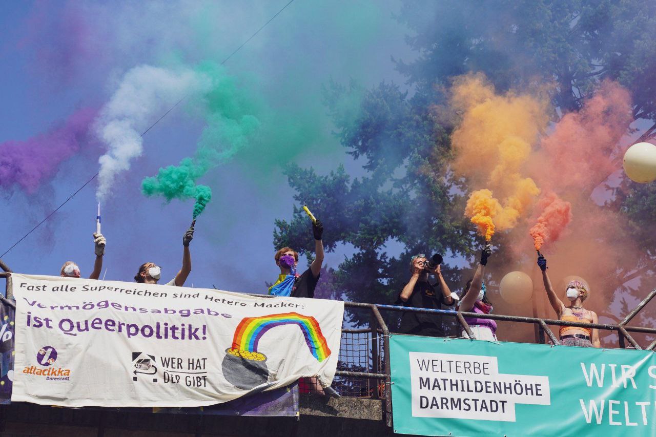 An einer Brücke hängt ein Transparent mit der Aufschrift "Fast alle Superreichen sind Hetero-Macker: Vermögensabgabe ist queerpolitik" dahinter stehen Menschen die mehrere Rauchtöpfe halten, sodass die Farben einen Regenbogen ergeben