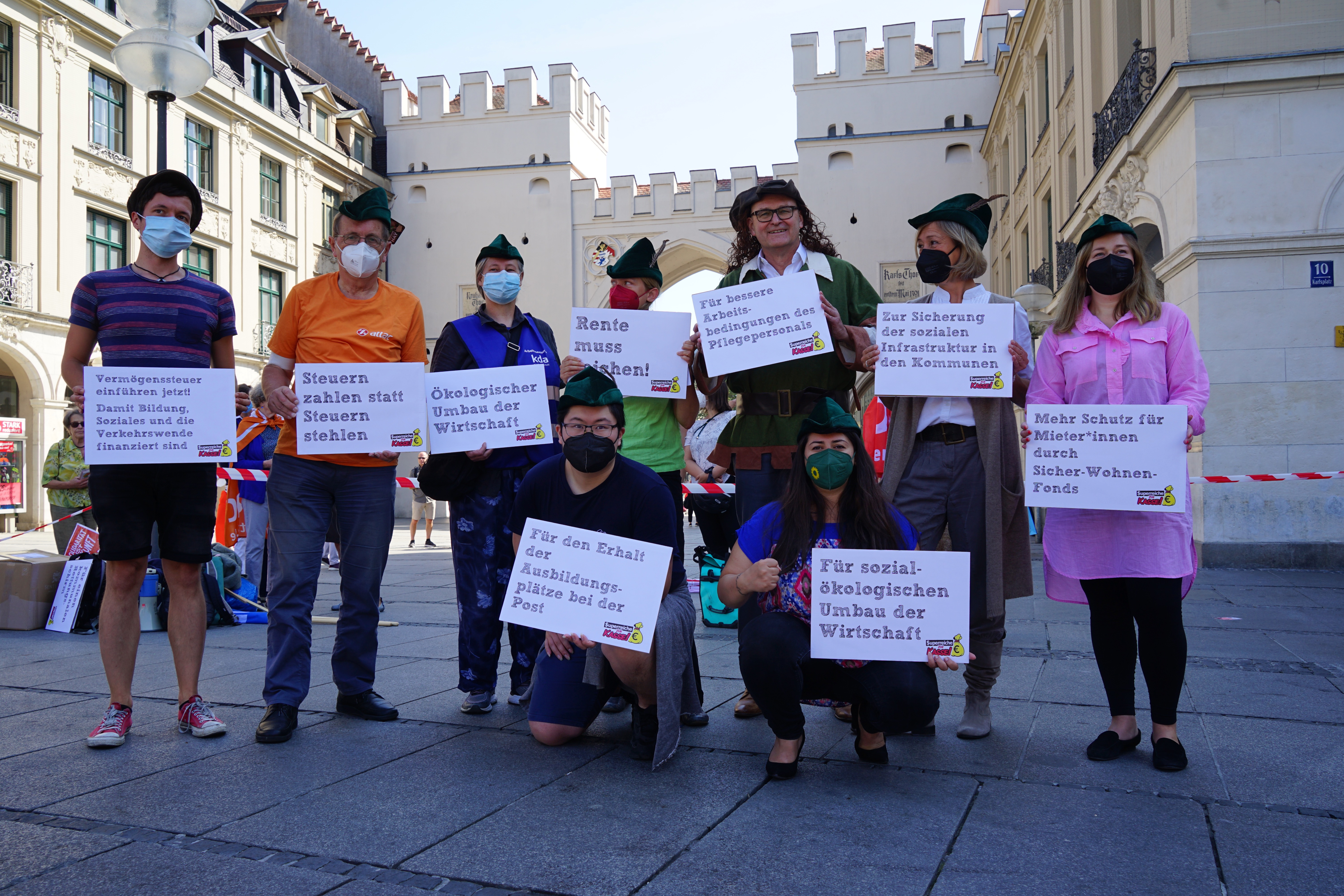 Gruppenbild des Bündnisses vorm Karlstor, die neun Teilnehmer*innen halten Schilder mit Umverteilungsforderungen in der Hand.