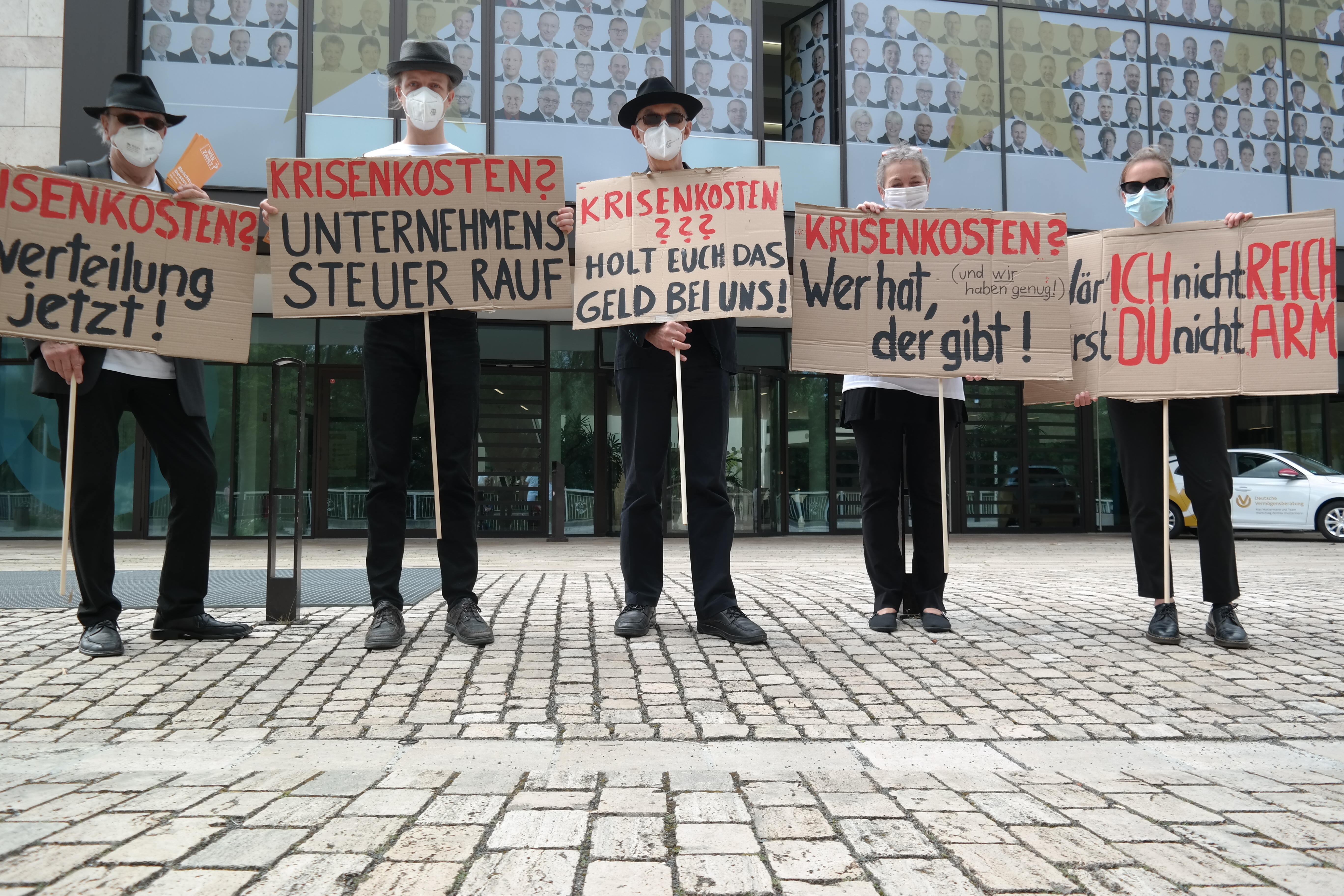 Eine Gruppe von Menschen in Anzügen steht vor der Zentrale der Deutschen Vermögensberatung mit Schildern auf denen Texte wie "Krisenkosten? Unternehmenssteuer Rauf" oder "Krisenkosten? Wer hat der gibt!" steht