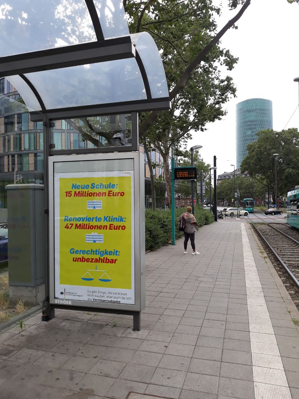 Im Bild ist ein Plakat der fiktiven Regierungskampagne im Schaukasten einer Straßenbahnhaltestelle, das Plakat trägt den text "Neue Schule: 15 Millionen Euro. Renoviertes Krankenhaus: 47 Millionen Euro. Gerechtigkeit: unbezahlbar. Es gibt Dinge, die kann man nicht kaufen, aber auch da hilft die Vermögensabgabe.