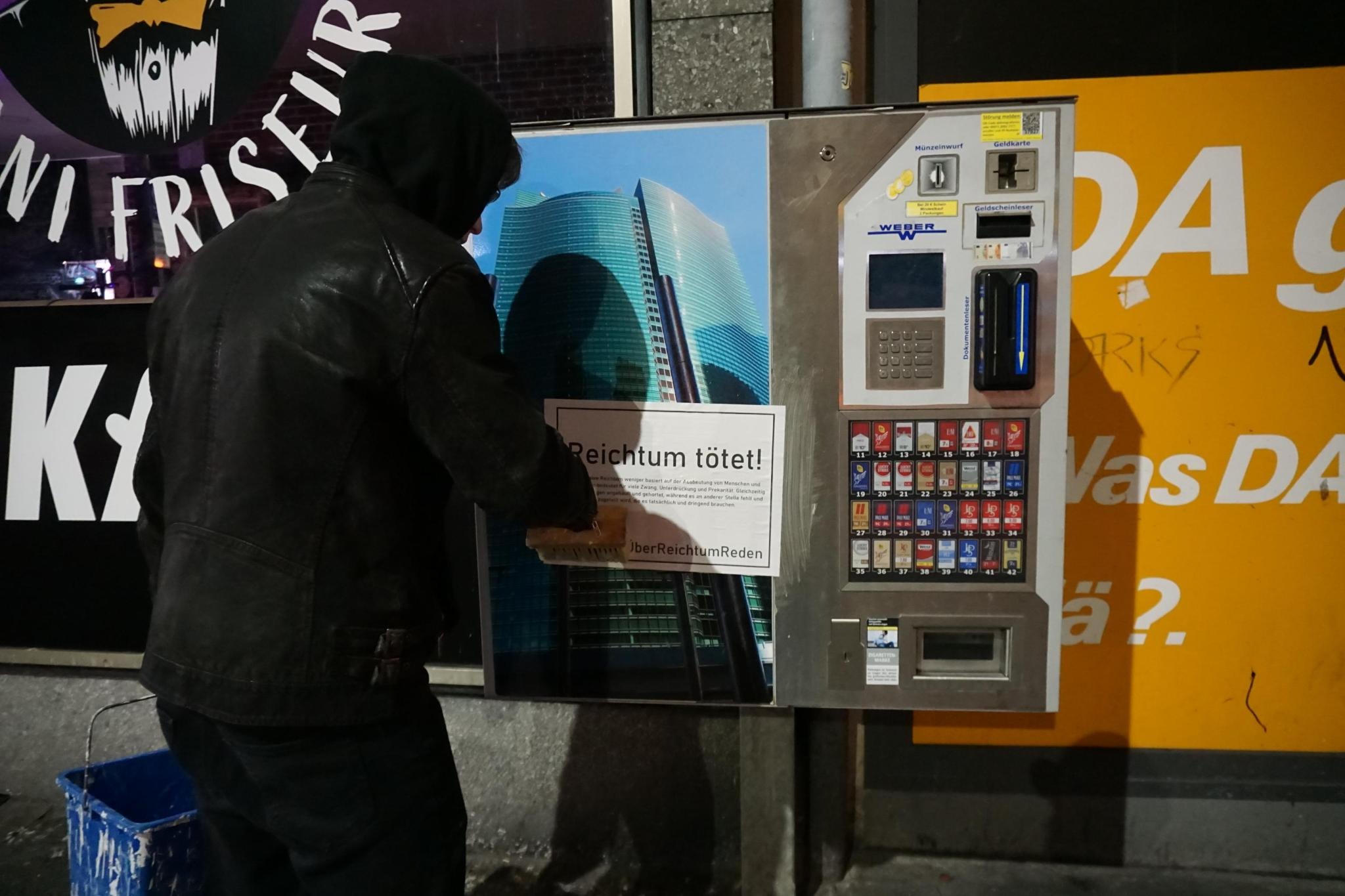 Ein schwarz gekleideter Aktivist klebt ein Plakat mit dem Slogan "Reichtum tötet" an einen Zigarettenautomaten.