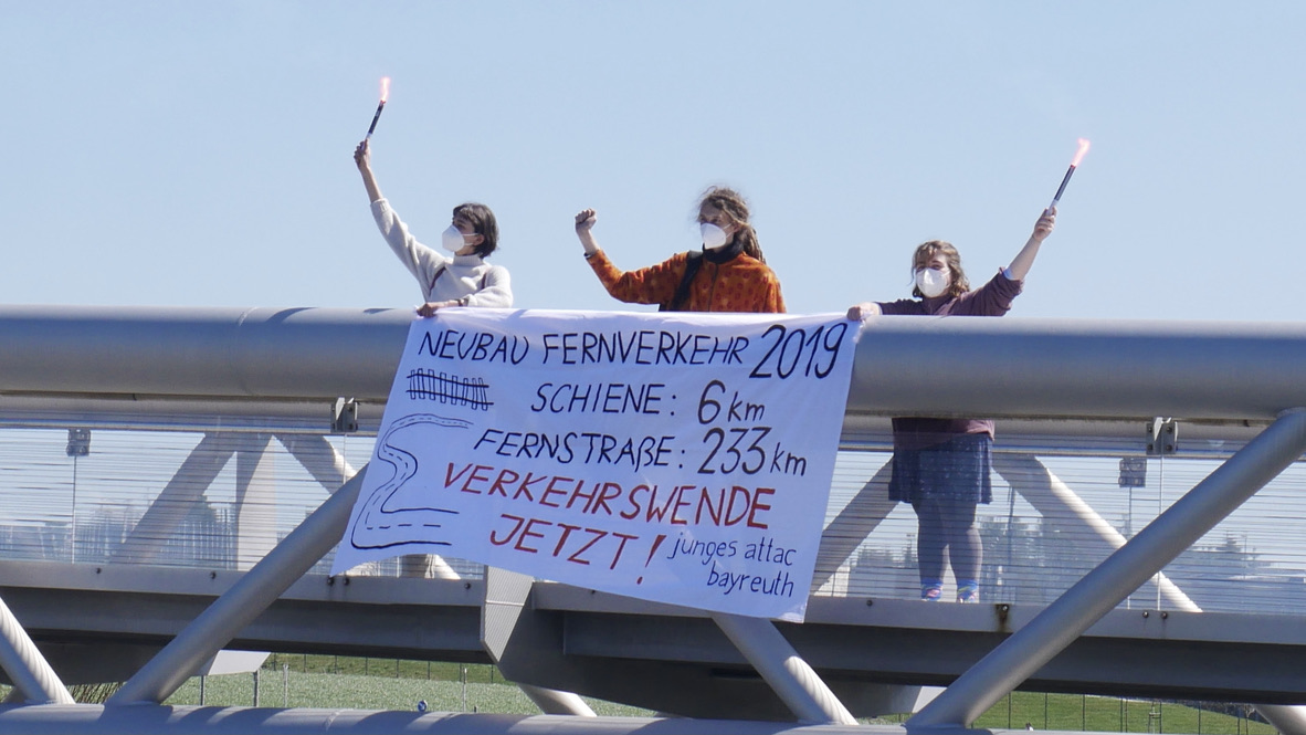 junge Attacis stehen auf einer Brücke mit Pyrotechnik in der Hand und halten ein Transparent mit der Aufschrift "Verkehrswende jetzt" und einem Vergleich zwischen dem Schienen- und dem Autobahnausbau in Deutschland