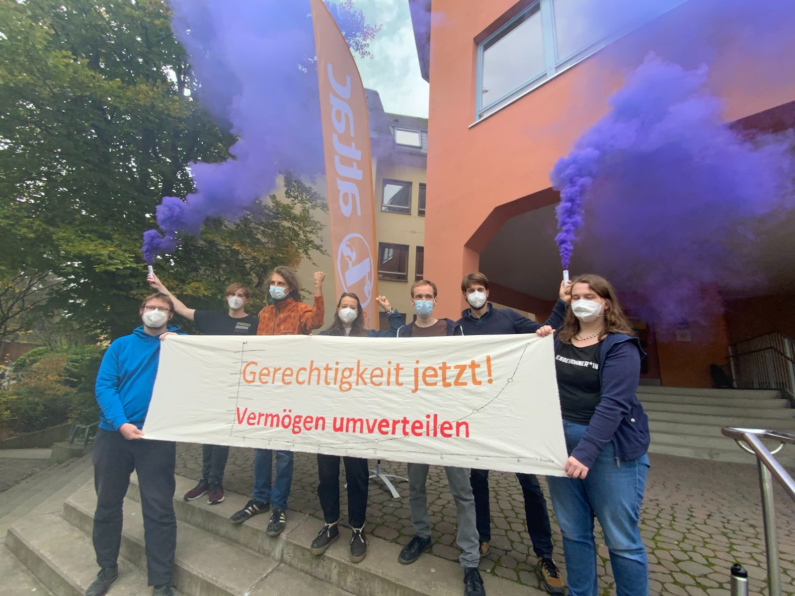 Junge Attacies stehen auf einer Treppe und halten ein Transparent mit der Aufschrift "Gerechtigkeit jetzt – Vermögen besteuern" und Lila Rauch.