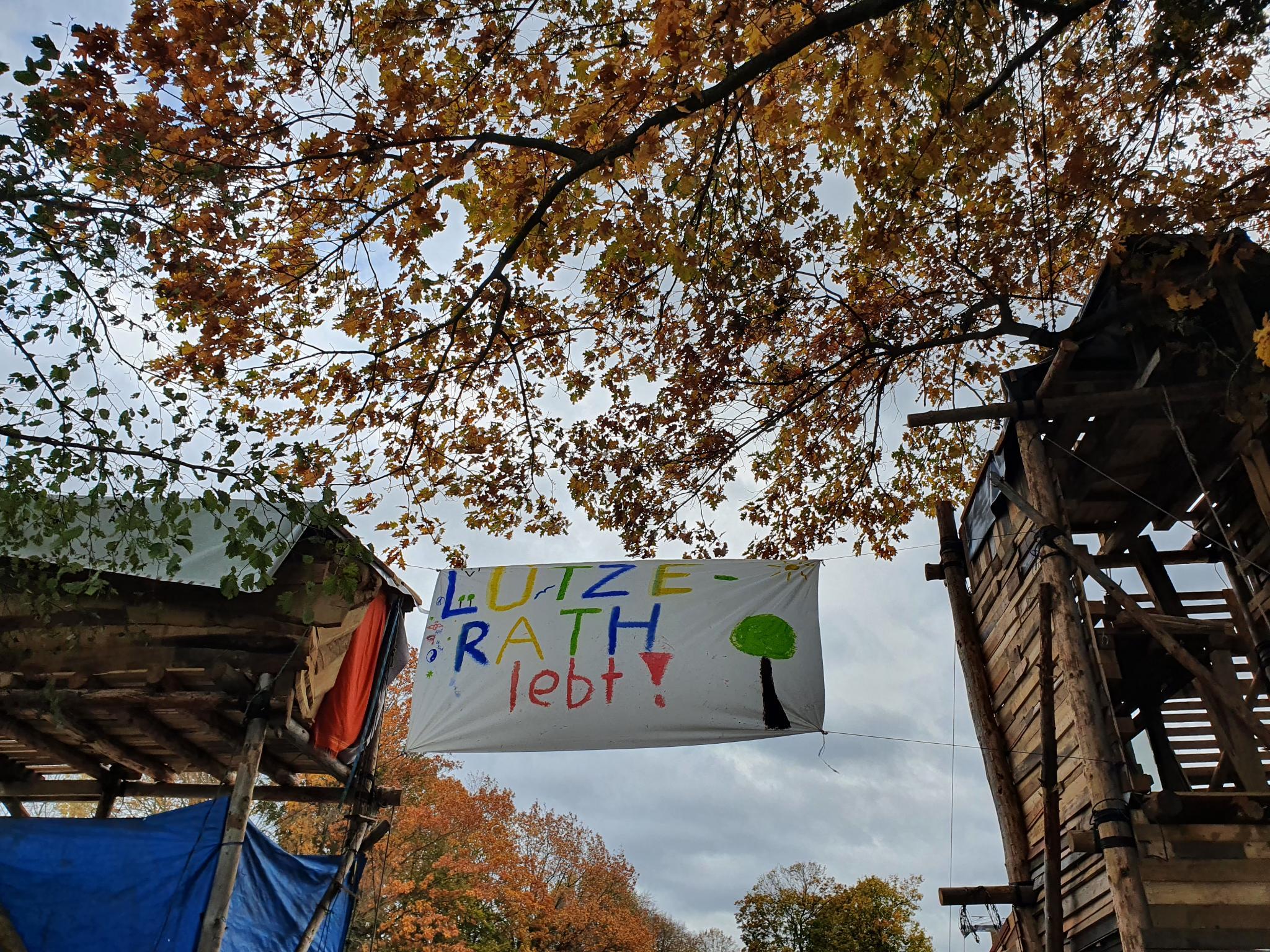 Zwischen zwei Protestcamphütten ist ein Transparent mit dem Slogan "Lützerath lebt!" aufgespannt.